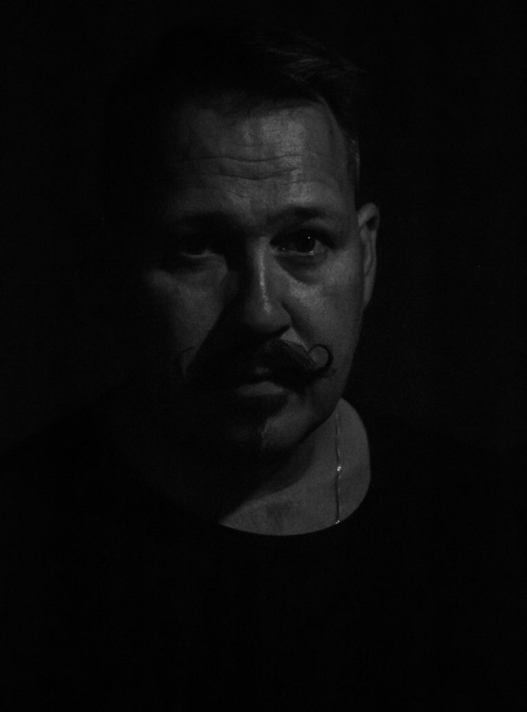 Schwarz-Weiß-Charakterporträt eines Mannes mit Moustache – geheimnisvolle Licht-Schatten-Wirkung, dunkler Hintergrund, intensiver Ausdruck.