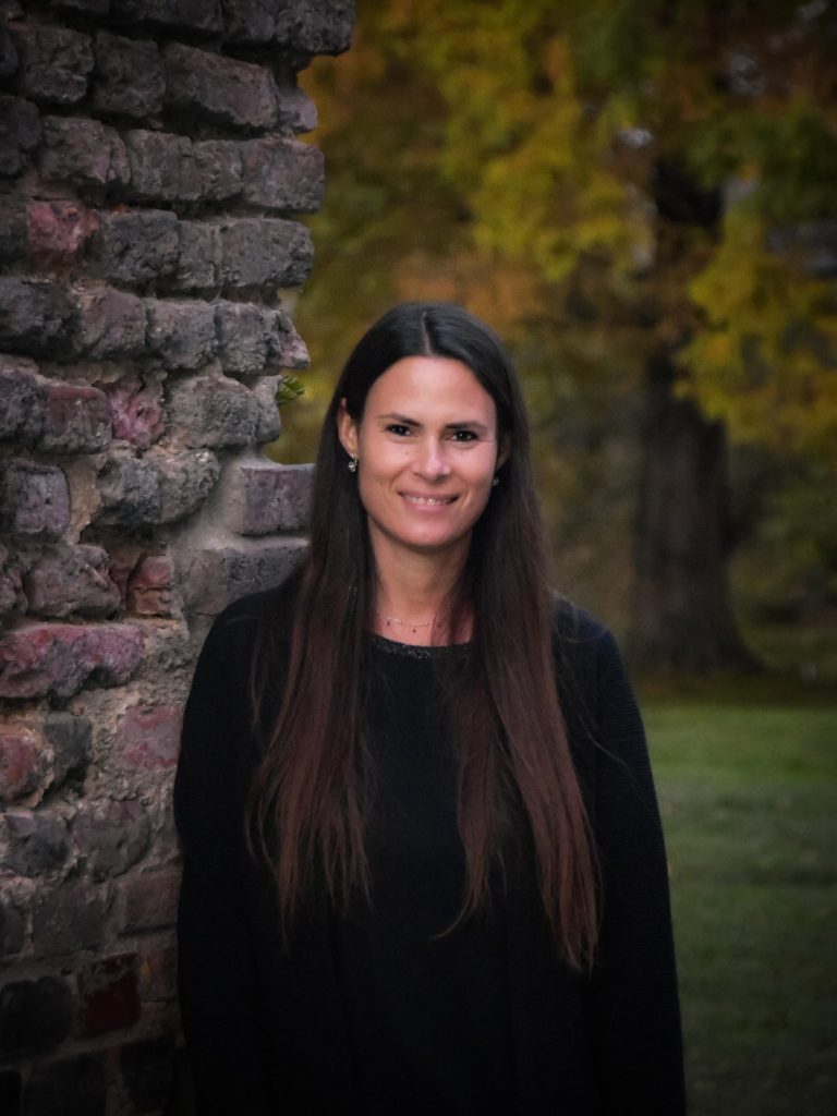 Natürliches Porträt einer schönen Frau im Herbst – sie lehn an einer alten Burgmauer, im Hintergrund verschwommen der Schlosspark.
