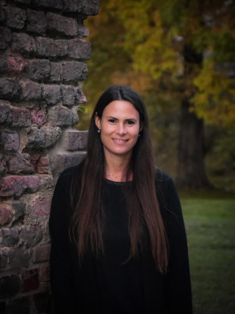 Natürliches Porträt einer schönen Frau im Herbst – sie lehn an einer alten Burgmauer, im Hintergrund verschwommen der Schlosspark.