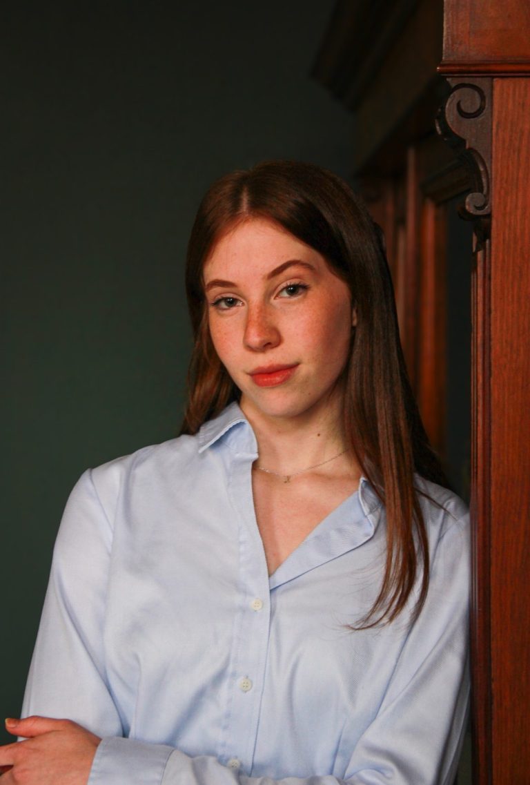 Klassische Eleganz – Bewerbungsfoto mit zeitloser Ausstrahlung Stilvolles Bewerbungsfoto – elegante Frau, lässig an einem antiken Schrank lehnend, dunkler Hintergrund.